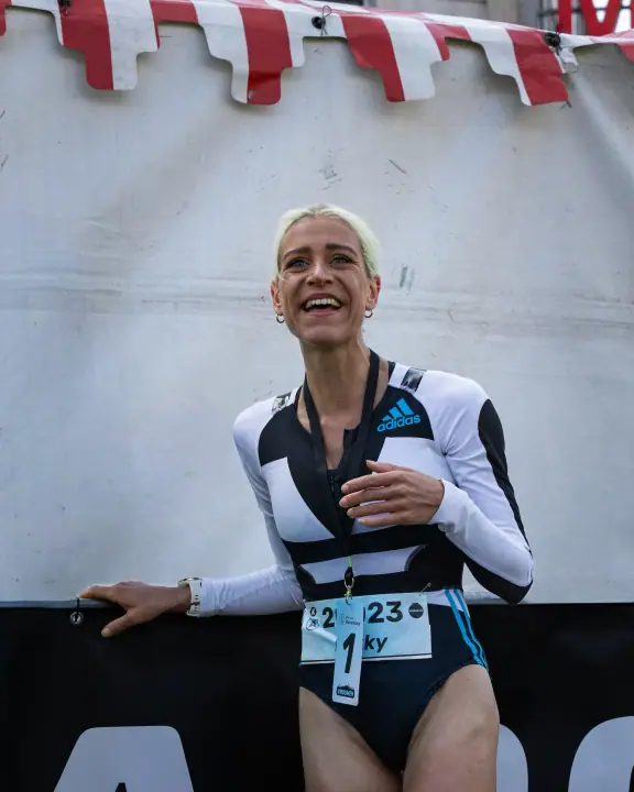 Lachende winnares met startnummer 1 leunt na haar finish ontspannen tegen een dranghek