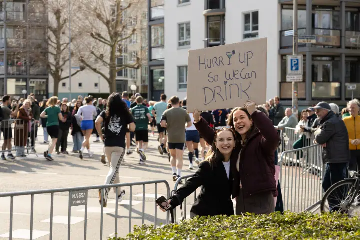 Twee vrouwen houden lachend een bordje met een grap omhoog voor de passerende lopers