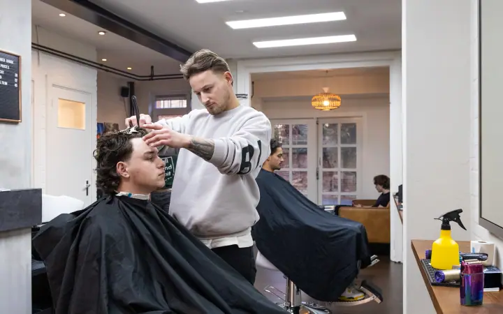 Barbier knipt krullend haar met schaar en kam terwijl het achterin de zaak ook druk is