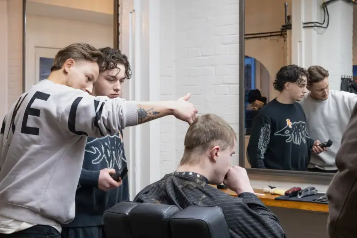 Barbier wijst een collega op een detail in het kapsel van een klant, gespiegeld in de wandspiegel