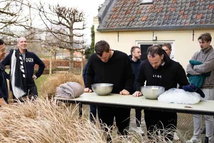 Brede opname van de ijswateroefening in de tuin, met siergras op de voorgrond en kijkende groepsgenoten.