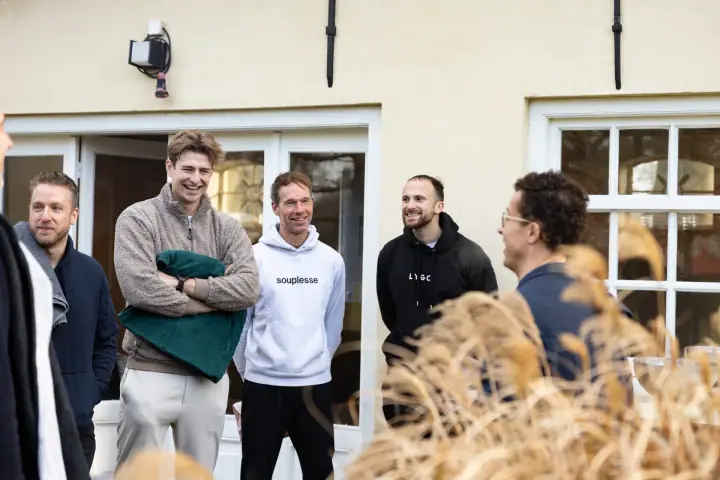 Vier deelnemers glimlachen naar de coach, die half van de camera afgedraaid achter het siergras staat.