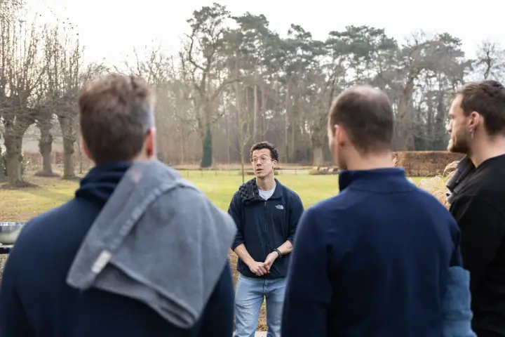 De coach spreekt de groep toe op het gazon, gefotografeerd over de schouders van de luisteraars.