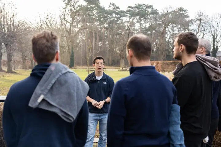 Deelnemers met een handdoek over de schouder luisteren in de tuin naar de uitleg van de coach.