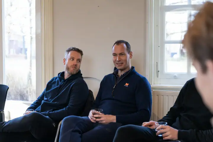 Twee deelnemers zitten binnen bij het raam met een glas in de hand en luisteren aandachtig.