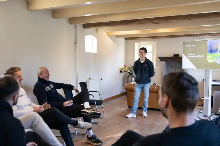 De coach lacht voor de groep in een zaal met houten balken, op het scherm staat workshop adem en ijsbad.