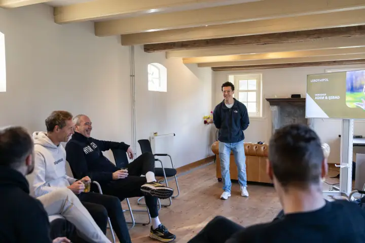 Deelnemers zitten in een kring op stoelen en lachen mee met de coach tijdens het theoriedeel.