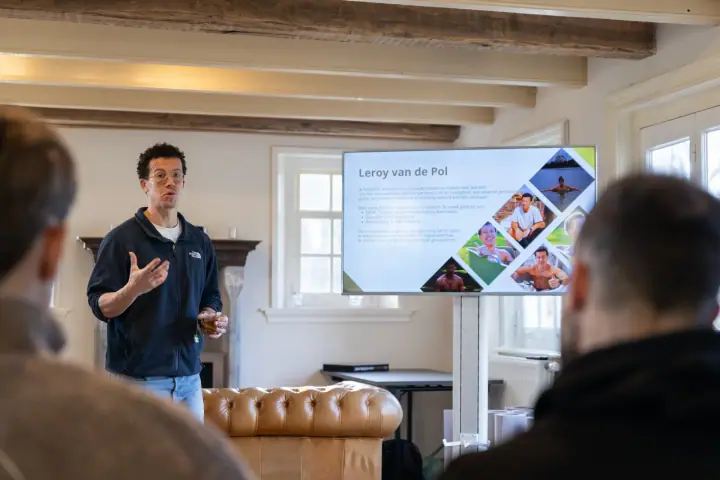 De coach presenteert naast een scherm met slides over zijn achtergrond, voor een leren chesterfieldbank.
