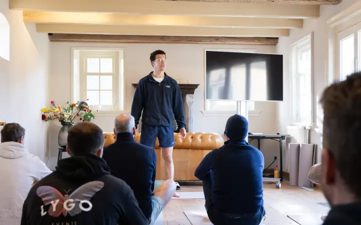 De coach in korte broek staat voor de groep, die op matjes op de houten vloer zit.