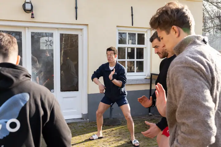 Buiten doet de coach een houding voor en de deelnemers bootsen hem na met geheven handen.