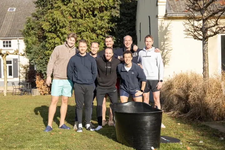 Groepsfoto van de deelnemers en de coach op het gazon, met de zwarte ijsbadkuip vooraan.