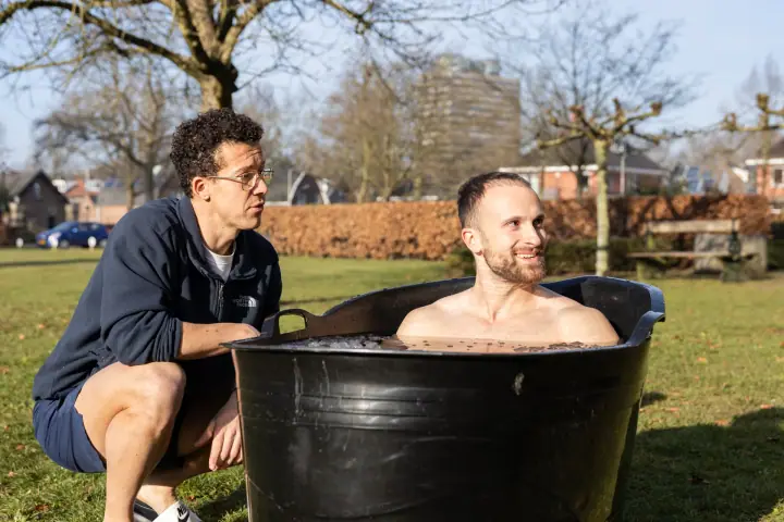 De coach zit gehurkt naast een glimlachende deelnemer die tot aan zijn schouders in het ijsbad zit.