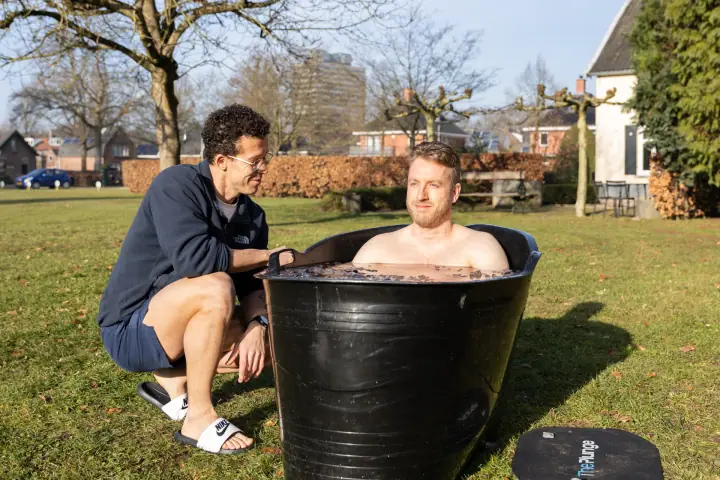 Een deelnemer zakt in de zwarte ijsbadkuip op het gras terwijl de coach hem rustig begeleidt.