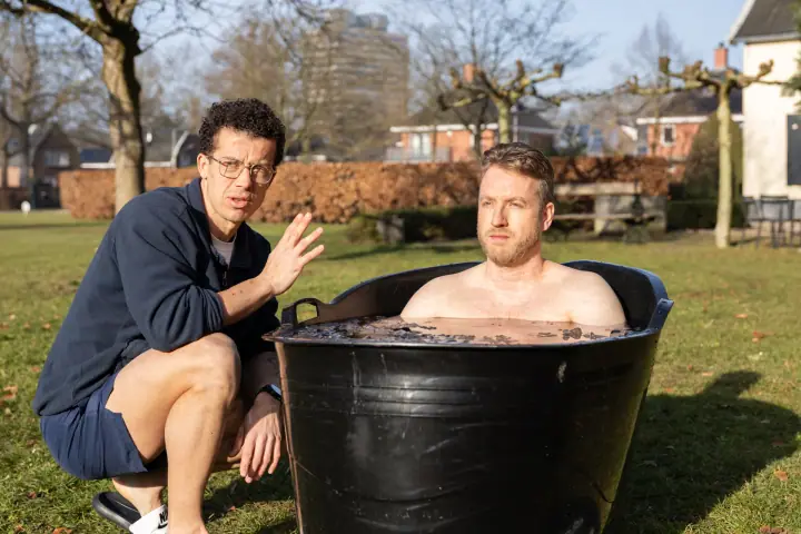 De coach gebaart rustig met zijn hand, de man in het ijsbad kijkt geconcentreerd voor zich uit.