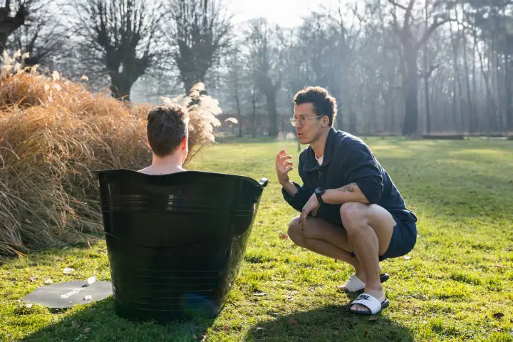 Tegenlicht in de tuin, de coach hurkt bij een deelnemer in het ijsbad en praat hem erdoorheen.