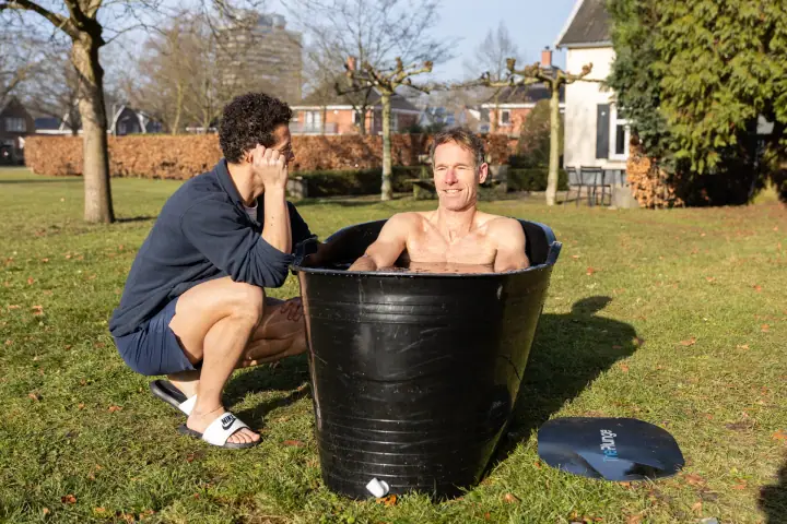 Een glimlach vanuit het ijsbad, terwijl de coach ernaast hurkt en met zijn hoofd in zijn hand luistert.