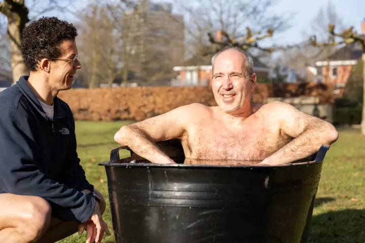 Lachende deelnemer met zijn armen over de rand van het ijsbad, de coach lacht met hem mee.