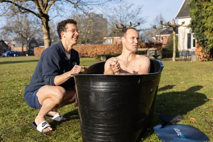 De coach lacht terwijl een deelnemer met gevouwen handen in de ijsbadkuip zit, op een zonnige winterdag.