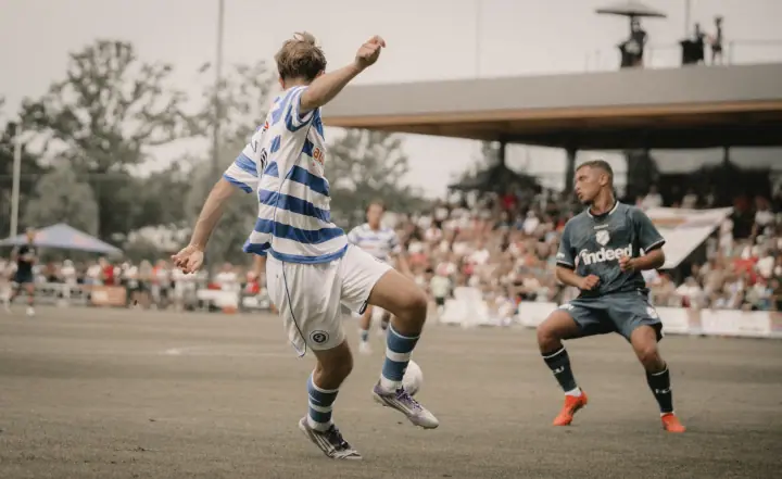 Jeugdspeler van De Graafschap haalt uit voor een volle tribune terwijl een verdediger in donker tenue blokt
