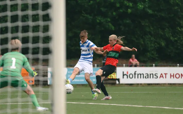 Aanvaller van De Graafschap schiet langs een NEC-verdediger, gezien door het net achter de keeper