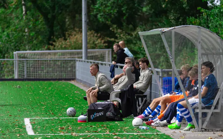 Staf en wisselspelers volgen de wedstrijd vanuit de dug-out langs het veld