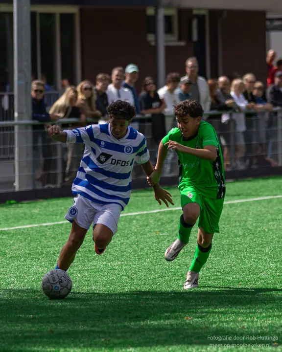Speler van De Graafschap dribbelt weg van een tegenstander in groen, publiek kijkt toe achter het hek