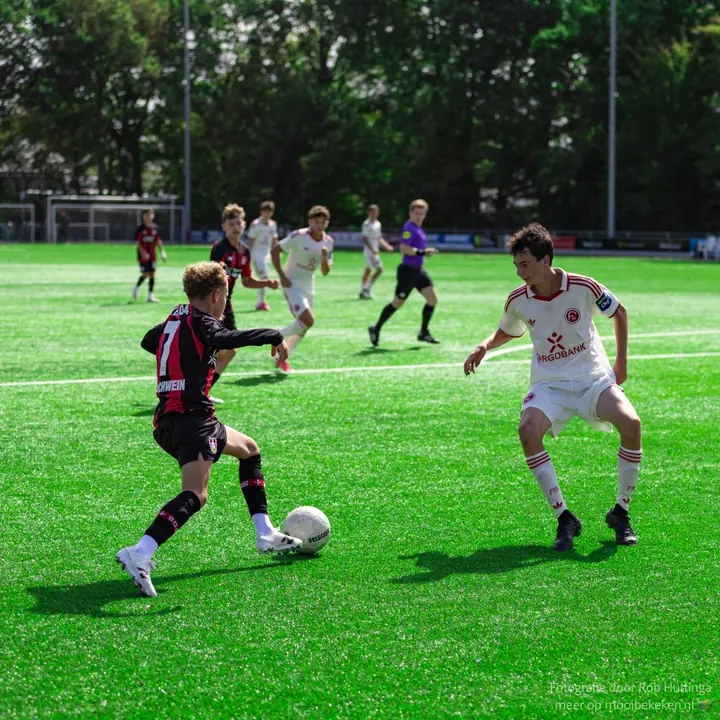 Speler in rood-zwart gestreept shirt dribbelt op een verdediger in wit tenue af