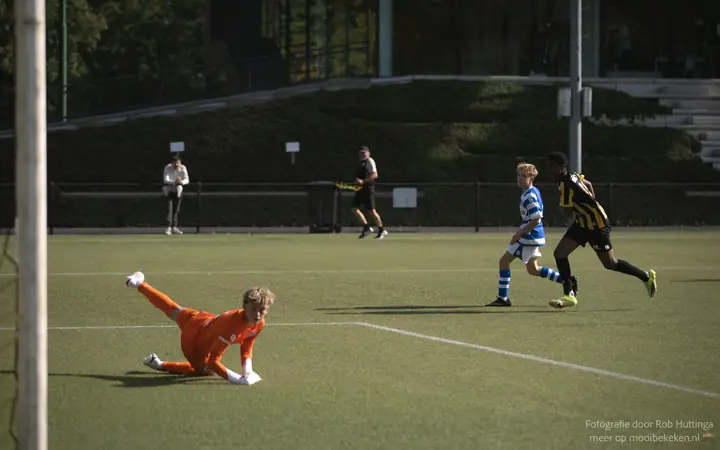 Keeper ligt languit op het gras terwijl twee spelers verderop doorlopen