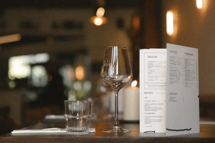 Gedekte tafel met wijnglas, waterglas en de kaart met snacks en bieren, sfeerlicht van Jadda erachter