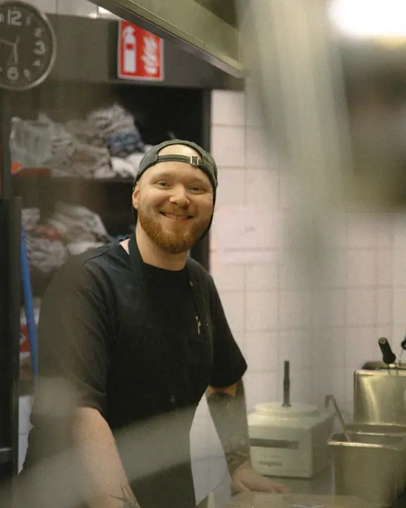 Glimlachende kok met baard en achterstevoren pet in de keuken, recht in de camera kijkend