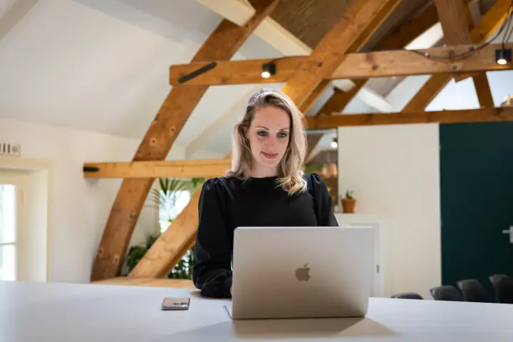 Blonde vrouw achter haar laptop onder de houten balken van een licht zolderkantoor