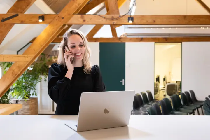 Lachend aan de telefoon achter de laptop, in een zolderkantoor met houten spanten en een groene deur
