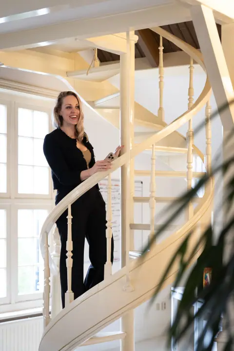 Lachend op de witte wenteltrap met telefoon in de hand, een plant onscherp op de voorgrond