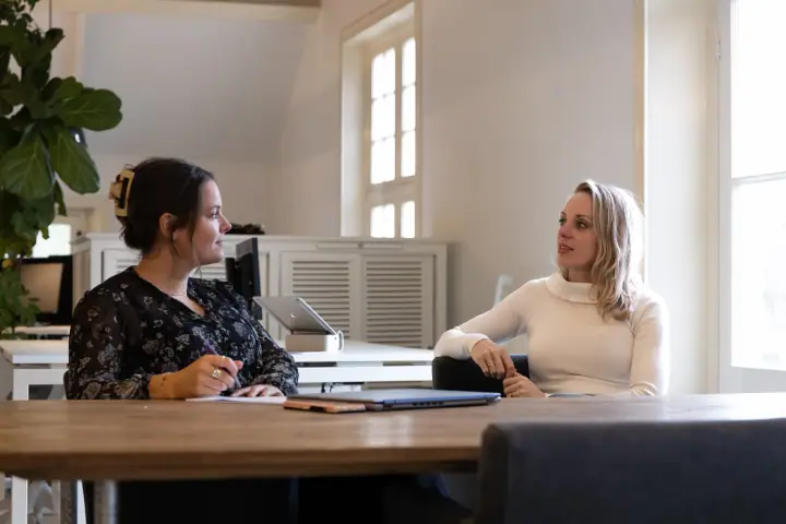 Twee vrouwen in gesprek aan een houten tafel in een licht kantoor met hoge ramen