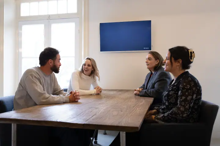 Vier mensen in gesprek aan een lange tafel, de blonde vrouw lacht naar de man met baard