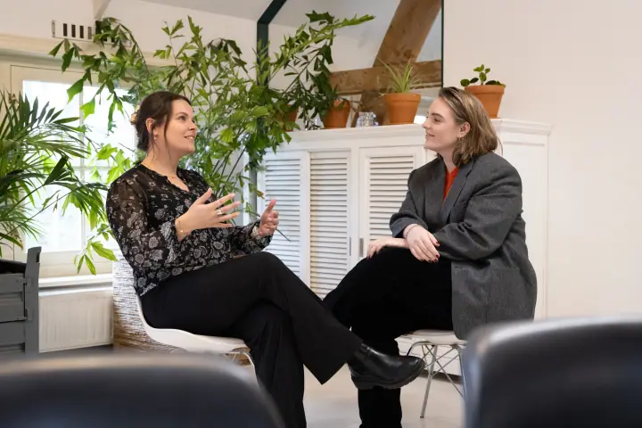 Twee vrouwen in gesprek op witte stoelen tussen de kamerplanten, een van hen gebaart