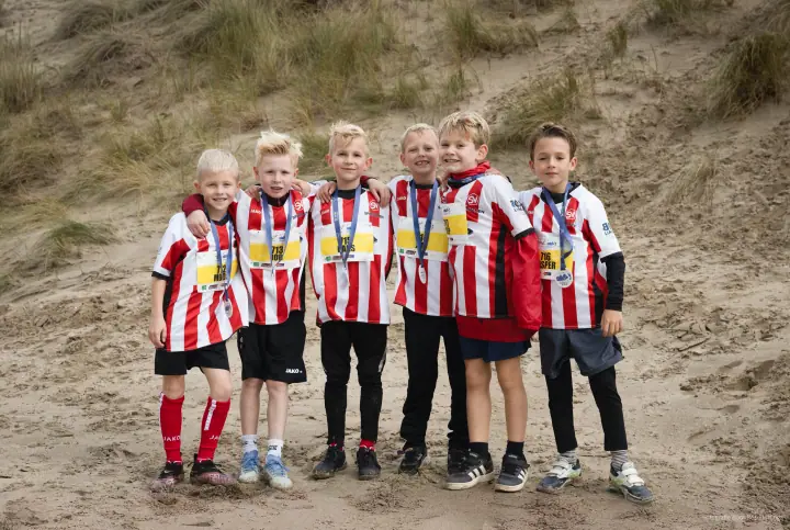Zes jongens in rood-wit gestreepte voetbalshirts met medaille en startnummer, arm in arm voor een duin.