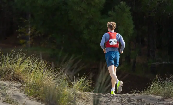 Trailrunner met rood hesje en startnummer loopt een zandpad in de duinen op, richting de dennen.