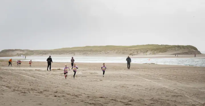 Kinderen in gestreepte shirts rennen over het brede strand bij Mudsweattrail 2025, ouders lopen mee.