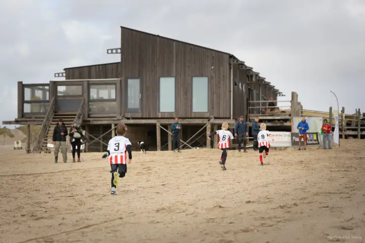 Drie jonge lopers met rugnummer 3, 6 en 8 sprinten over het zand naar een houten strandpaviljoen.