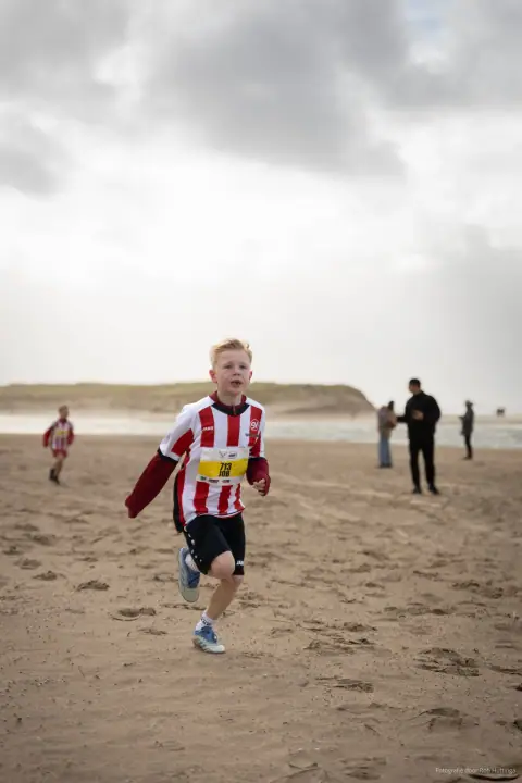 Jongen in rood-wit shirt met startnummer rent over het strand op de camera af, onder een grijze lucht.