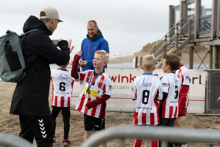 Een jonge loper geeft een high five aan een man met een telefoon, teamgenoten in gestreepte shirts erachter.