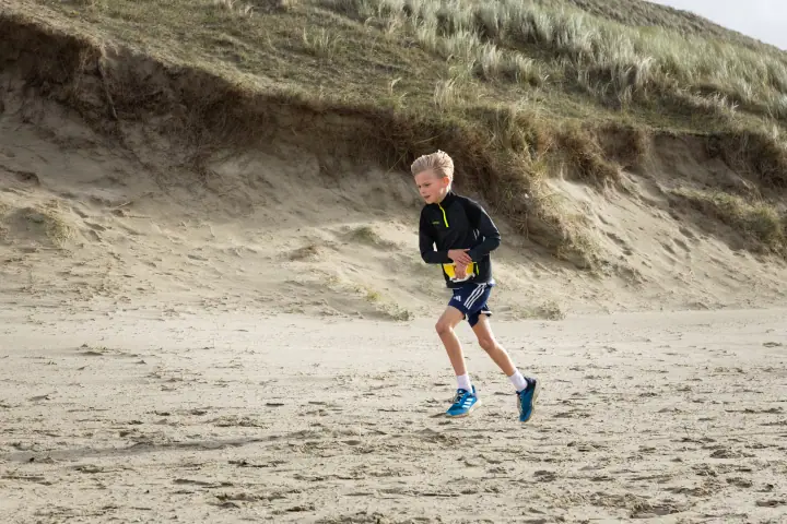 Jongen in zwart jack rent over het strand langs de voet van een steile duinrand.