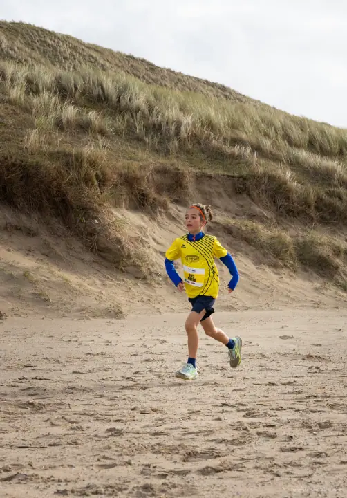 Meisje in geel shirt met startnummer loopt soepel over het zand onder een begroeide duinhelling.
