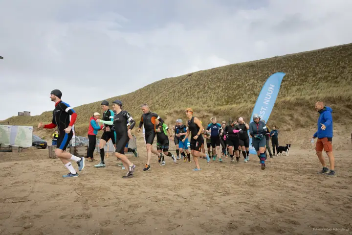 Start van MudSweatTrail op het strand, lopers in wetsuits vertrekken met de duinen achter zich.