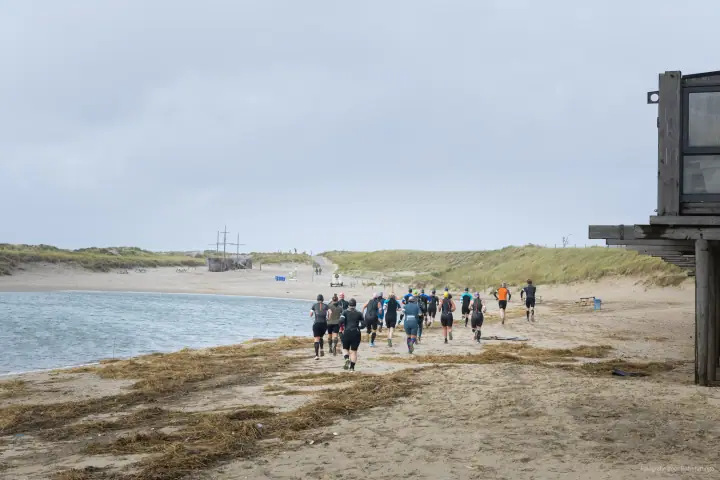 Een groep lopers in wetsuits trekt langs de waterlijn het strand af, richting de duinen.