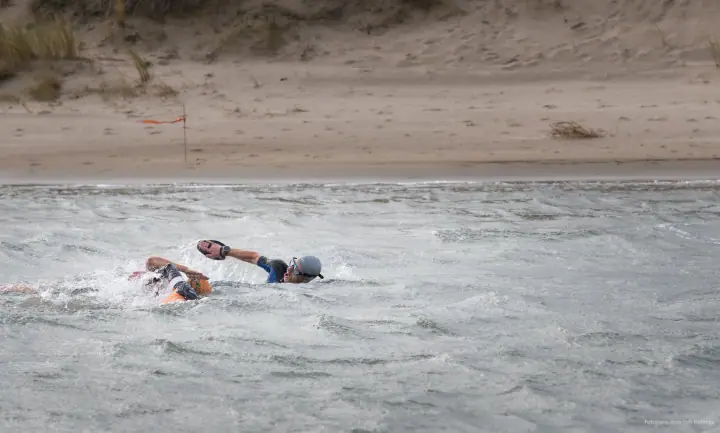Twee zwemmers in wetsuits ploeteren door woelig water, een van hen met handpeddels.