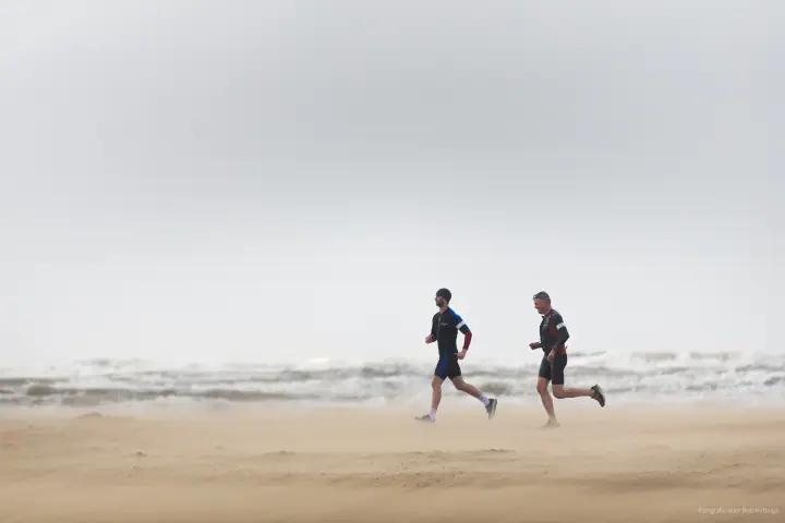 Twee lopers in triatlonpakken rennen over het strand terwijl het zand om hun benen stuift.