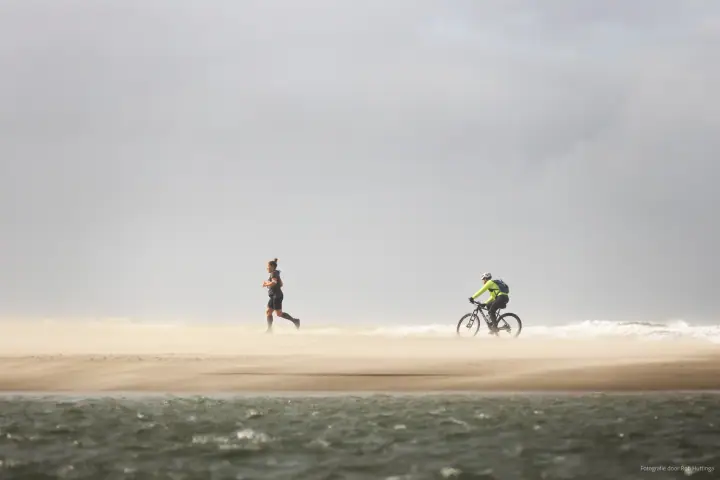 Een loper en een mountainbiker steken het strand over in een wolk opwaaiend zand.