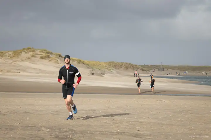 Loper in zwart pak met rode mouwen komt over het zandstrand aangelopen, duinen en donkere lucht erachter.
